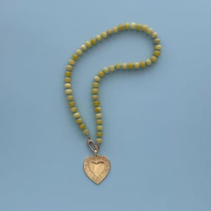 May include: A beaded necklace with a gold heart pendant. The necklace features light green and yellow beads. The heart pendant has a textured surface and a smaller heart shape in the center. The necklace is displayed on a light blue background.