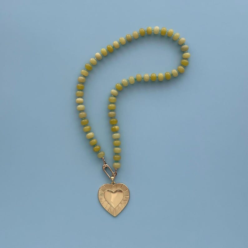May include: A beaded necklace with a gold heart pendant. The necklace features light green and yellow beads. The heart pendant has a textured surface and a smaller heart shape in the center. The necklace is displayed on a light blue background.