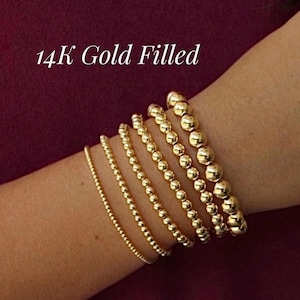 May include: A set of six gold-filled beaded bracelets featuring varying bead sizes arranged in a row. The bracelets are made of polished, reflective gold-colored spherical beads. The text 14K Gold Filled is displayed in white above the bracelets.