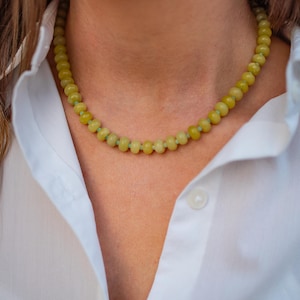 May include: A beaded necklace with yellow-green beads. The necklace is worn over a white button-down shirt. The beads are round and evenly spaced, creating a simple yet elegant design. The necklace is a natural accessory.