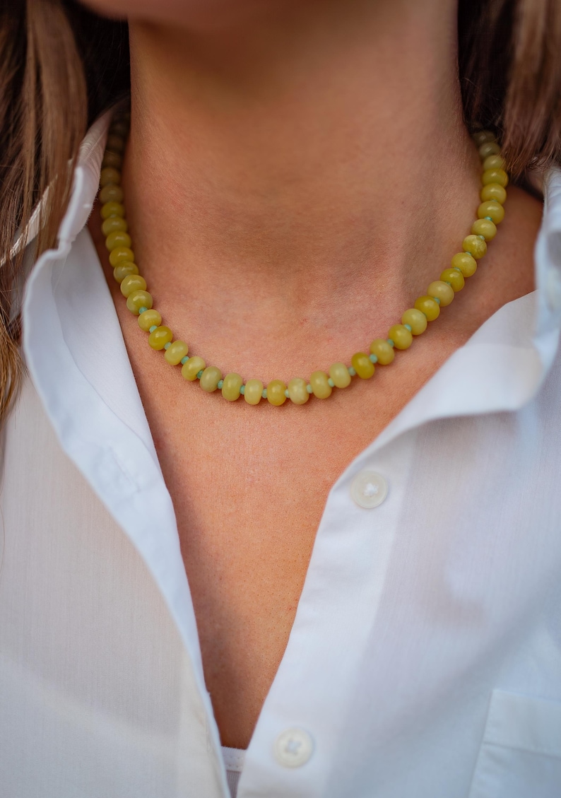 May include: A beaded necklace with yellow-green beads. The necklace is worn over a white button-down shirt. The beads are round and evenly spaced, creating a simple yet elegant design. The necklace is a natural accessory.
