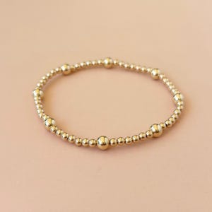 May include: Gold beaded bracelet with varying sized round beads. The bracelet is made of small, shiny, gold-colored beads strung together, with larger beads interspersed throughout. The bracelet is displayed on a light pink background.