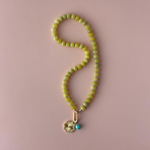 May include: A yellow beaded necklace with a gold pendant and a small turquoise bead. The pendant is a gold flower shape. The necklace is on a pink background.