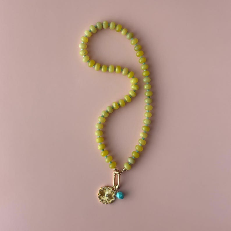 May include: A yellow beaded necklace with a gold pendant and a small turquoise bead. The pendant is a gold flower shape. The necklace is on a pink background.