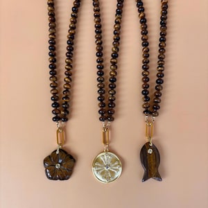 May include: Three necklaces featuring brown and black tiger eye beads strung on cord. Each necklace includes a gold-toned metal link connecting to a pendant: one carved tiger eye flower, one round gold-toned medallion with crystals, and one fish-shaped tiger eye.