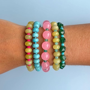 May include: A set of five beaded bracelets featuring various colors and textures. One bracelet has yellow beads with bright pink spacers, another has turquoise blue beads, a third has pink beads with small green spacers, a fourth has yellow beads with small green spacers, and the last has faceted dark green beads with turquoise blue spacers.