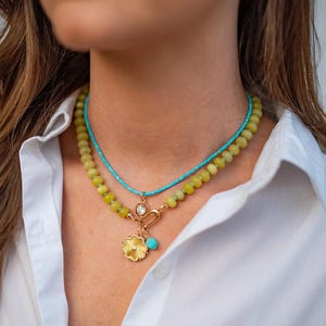 May include: A layered necklace featuring a turquoise beaded strand and a strand of yellow-green beads. The lower necklace has a gold pendant with a flower design and a turquoise charm. A gold clasp and a clear gem are also visible.