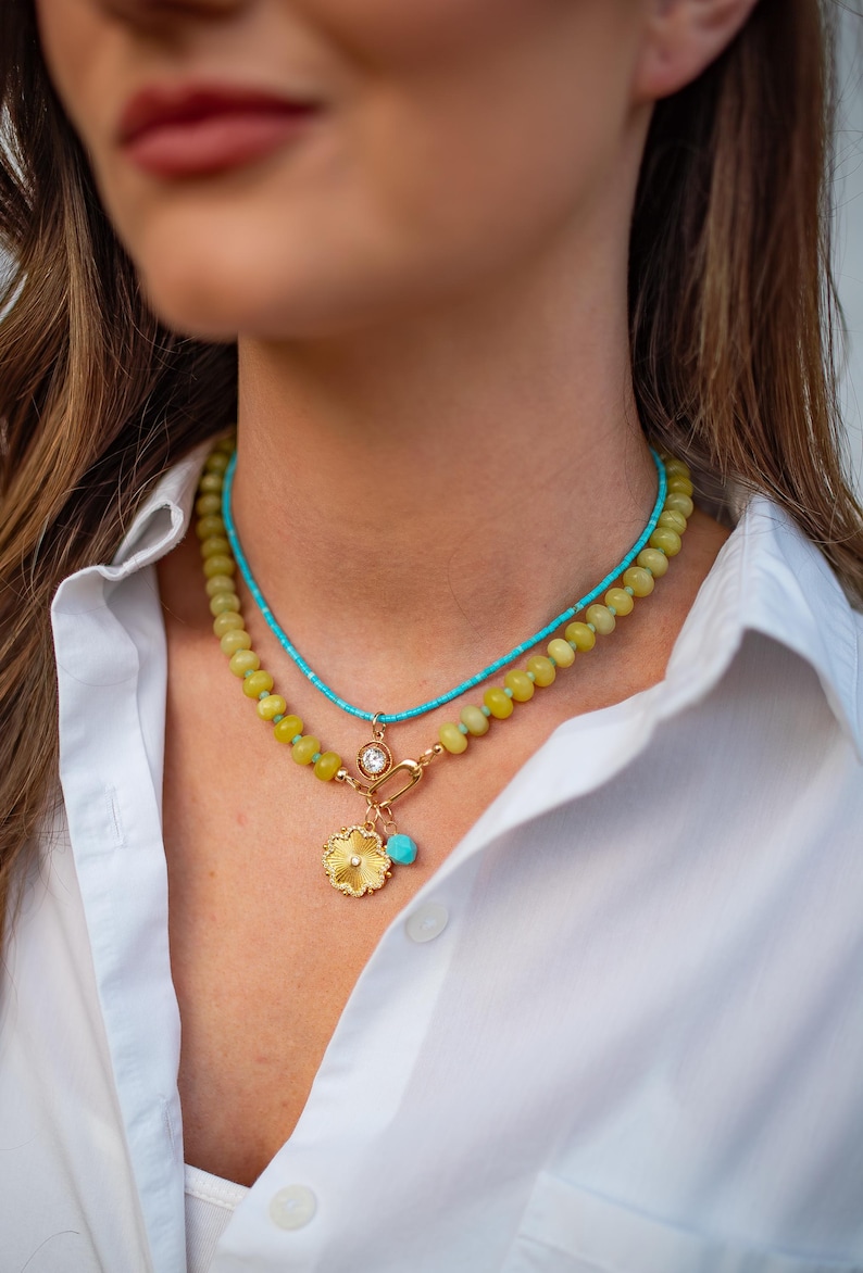 May include: A layered necklace featuring a turquoise beaded strand and a strand of yellow-green beads. The lower necklace has a gold pendant with a flower design and a turquoise charm. A gold clasp and a clear gem are also visible.