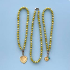 May include: Three necklaces with yellow and cream-colored beads. One necklace has a gold heart pendant, another has a gold flower pendant with a small turquoise bead, and the third necklace has a gold clasp. The necklaces are displayed on a light blue background.