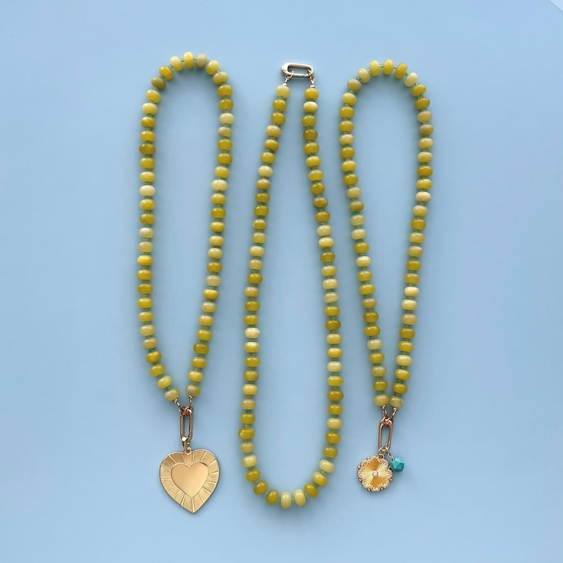 May include: Three necklaces with yellow and cream-colored beads. One necklace has a gold heart pendant, another has a gold flower pendant with a small turquoise bead, and the third necklace has a gold clasp. The necklaces are displayed on a light blue background.