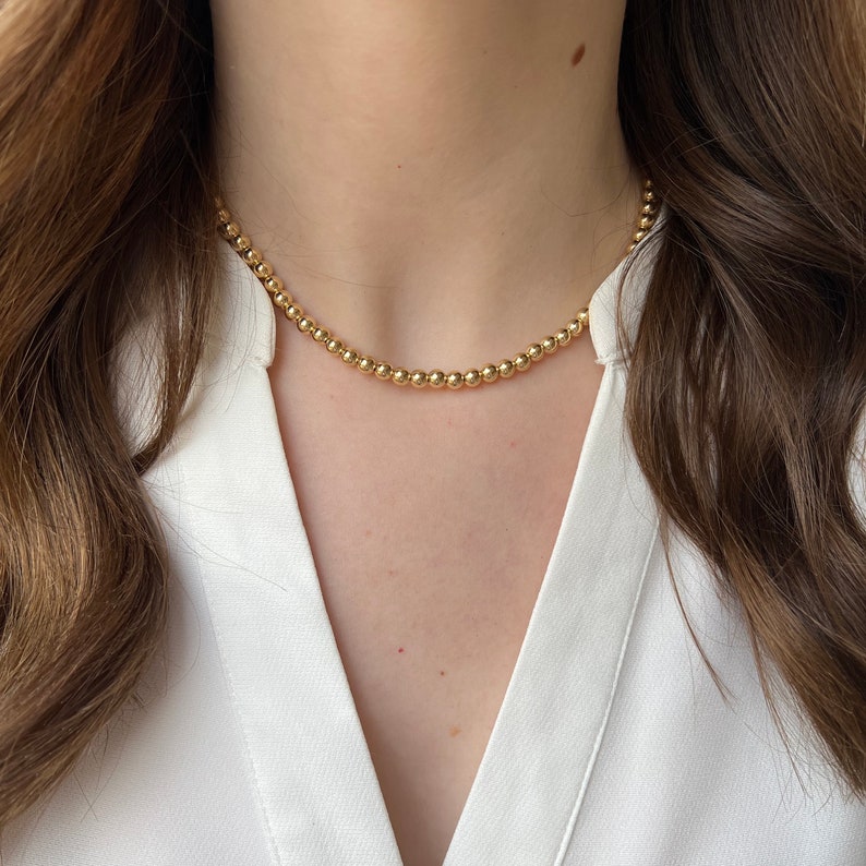 May include: A gold beaded necklace with a delicate chain. The necklace is worn by a person with long brown hair and a white shirt.