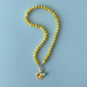 May include: A yellow beaded necklace with a gold-colored pendant in the shape of a flower. The pendant is connected to a gold-colored clasp, and a small turquoise bead hangs below the pendant. The necklace is displayed on a light blue background.