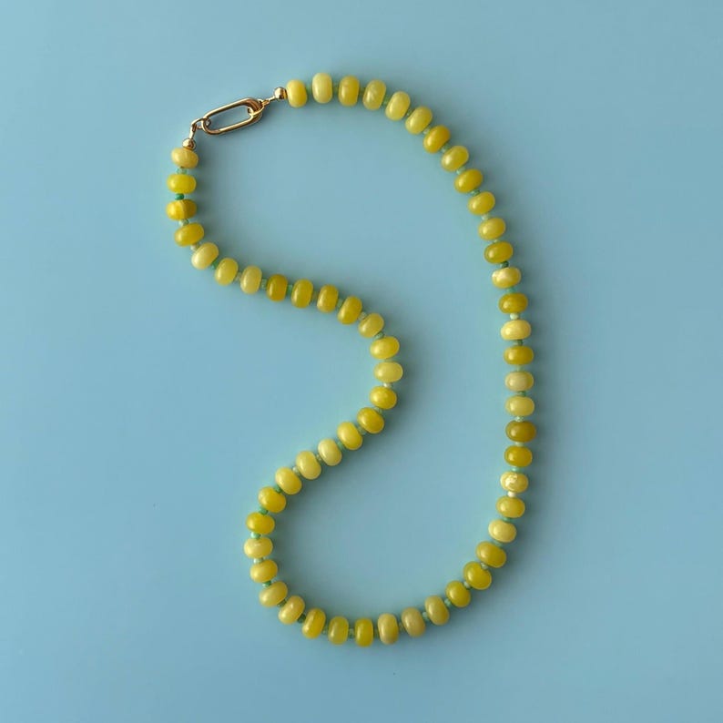 May include: A yellow beaded necklace with a gold clasp. The necklace is composed of small, rounded beads in varying shades of yellow, creating a gradient effect. The clasp is a simple, elegant design. The necklace is displayed against a light blue background.
