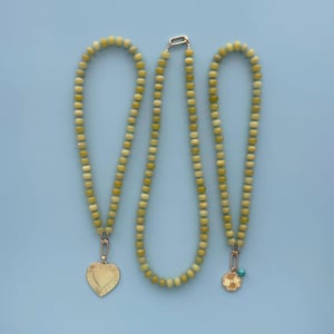 May include: Three necklaces with light green and yellow beads are displayed against a light blue background. Two necklaces have gold-colored pendants, one heart-shaped and the other a flower with a turquoise accent. The third necklace is plain.