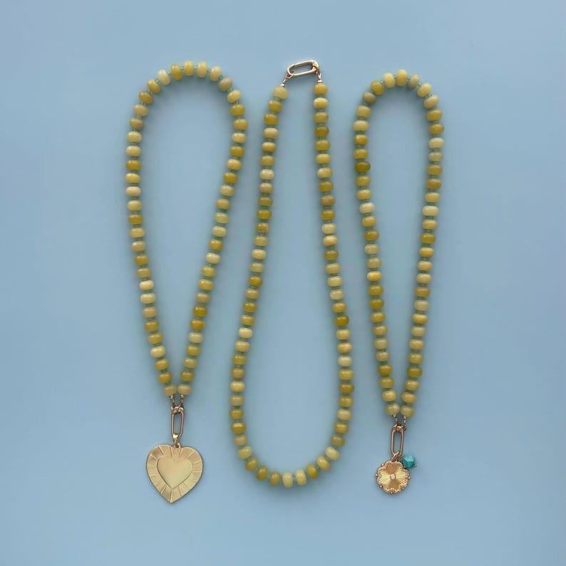 May include: Three necklaces with light green and yellow beads are displayed against a light blue background. Two necklaces have gold-colored pendants, one heart-shaped and the other a flower with a turquoise accent. The third necklace is plain.