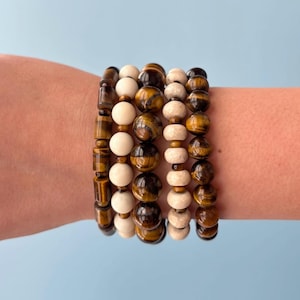May include: A set of five stacked bracelets featuring a mix of cylindrical and spherical beads. The design includes brown tiger eye stone beads with natural banding, smooth matte cream-colored beads, and textured light beige stone beads.