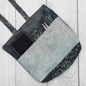 May include: A handmade tote bag with a black and gray floral and patterned design. The bag has a pocket with a gray dotted pattern and contains a smartphone and pens. The bag has two straps and is made of fabric.