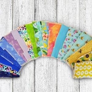 May include: A collection of 16 colorful fabric squares with various patterns, including butterflies, flowers, bees, and geometric designs. The squares are arranged in a semi-circle shape, with the yellow squares at the bottom.