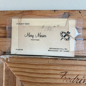 May include: Close-up of a framed artwork with a light-colored wooden frame. A label is attached to the back, displaying the artist's name, "Marg Messer PAINTINGS," and contact information. The frame has the name "Fredriv" carved into the wood.