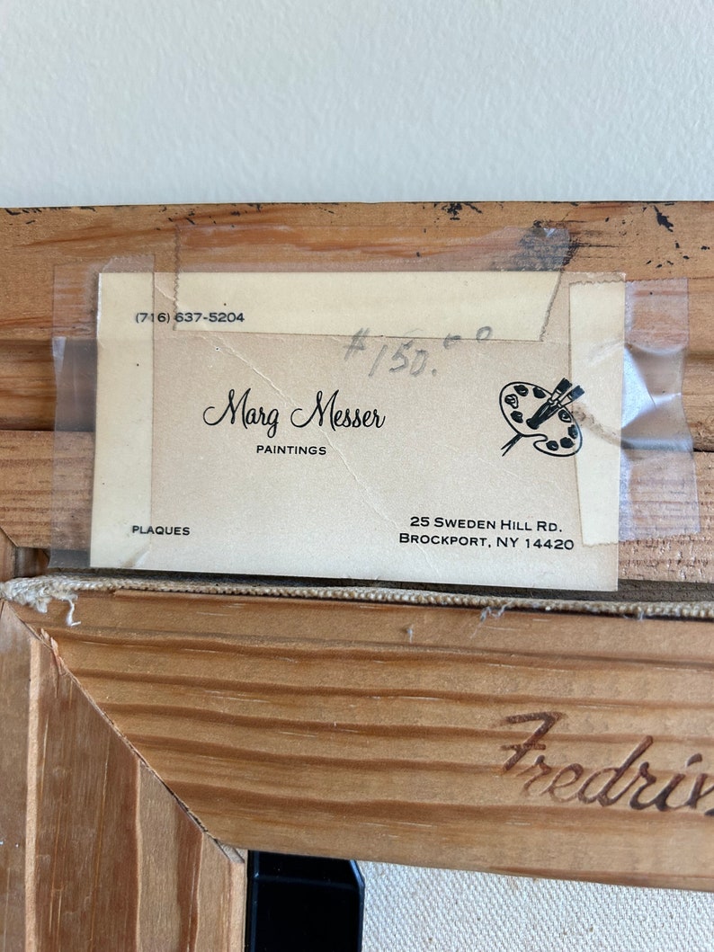 May include: Close-up of a framed artwork with a light-colored wooden frame. A label is attached to the back, displaying the artist's name, "Marg Messer PAINTINGS," and contact information. The frame has the name "Fredriv" carved into the wood.