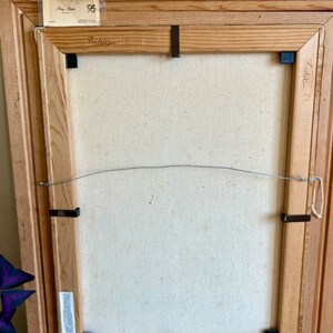 May include: Back of a framed painting with a light-colored canvas. The wooden frame is light brown and has a wire for hanging. The artist's name and date are visible on the frame. Black clips secure the canvas to the frame.