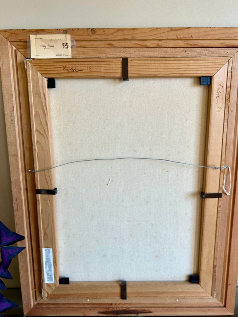 May include: Back of a framed painting with a light-colored canvas. The wooden frame is light brown and has a wire for hanging. The artist's name and date are visible on the frame. Black clips secure the canvas to the frame.