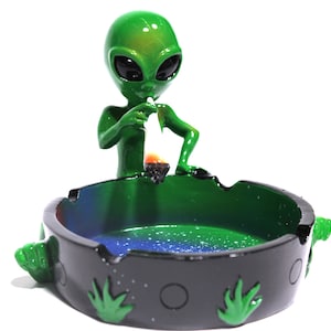May include: Green alien figurine smoking a pipe, sitting on a black and green ashtray with a weed leaf design. The ashtray has a blue and green interior.