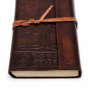 May include: A dark brown leather journal with a tree of life design embossed on the cover. The journal is secured with a leather strap. The edges are stitched with red thread. The journal is approximately 6 inches tall and 4 inches wide.