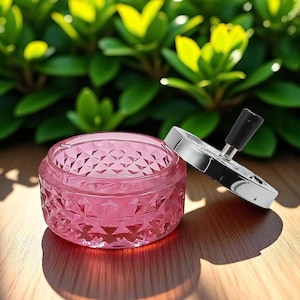 May include: A pink, diamond-patterned glass ashtray with a chrome and black lid sits on a wooden surface. The ashtray is open, casting a shadow. Green foliage is in the background.