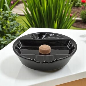 May include: A glossy black ceramic ashtray with a central compartment and a small, round, brown cork. The ashtray is on a white surface, with a blurred background of green plants and colorful flowers.