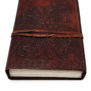 May include: A brown leather journal with an embossed floral design and a leather strap closure. The journal's cover features a detailed leaf pattern. The edges of the journal are decorated with a floral border. The journal is filled with blank pages.