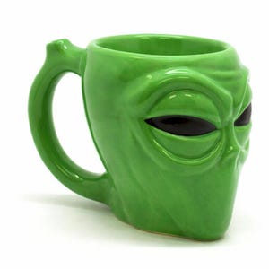 May include: A green ceramic mug shaped like an alien head with large black eyes. The mug has a curved handle and a glossy finish. The alien face is detailed with wrinkles and facial features. The mug is designed to hold beverages.