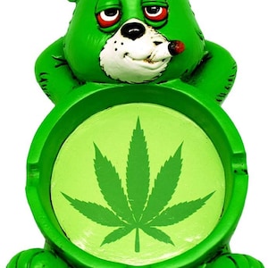 May include: Green ceramic ashtray shaped like a cartoon bear with a marijuana leaf design. The bear has red eyes and is holding a cigarette in its mouth.