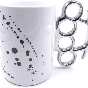 May include: White ceramic mug with a black and white splatter design and a silver metal knuckle duster handle.
