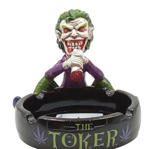 May include: A novelty ashtray featuring a sculpted figure of a character with green hair and a purple suit, holding a lit cigarette. The black ashtray has the words "THE TOKER" and cannabis leaf designs.