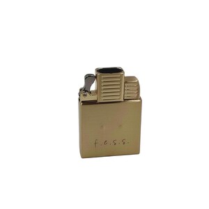 May include: A gold-colored lighter with a rectangular body and a hinged top. The lighter has a textured surface and the letters "f.e.s.s." are inscribed on the front.