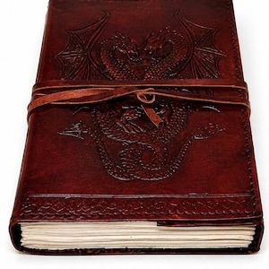 May include: A dark brown leather journal with a dragon embossed on the cover. The journal is secured with a leather strap. The dragon design features two dragons facing each other with wings spread. The journal's pages are visible.