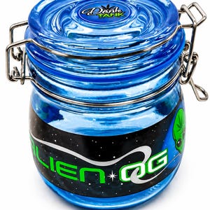 May include: A blue glass jar with a hinged lid and metal clasp. The jar has a black and blue label with the words "ALIEN OG" in green and white, along with an alien graphic. The lid has the words "DANK TANK" in white.