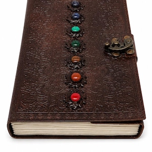 May include: A brown leather journal with embossed floral patterns and a metal clasp. The cover is adorned with a series of colorful gemstones, including red, orange, brown, green, blue, and purple. The journal is closed.