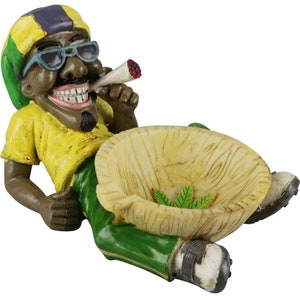 May include: A green, yellow, and purple figurine of a person smoking a joint, with a bowl-shaped ashtray in front of them. The ashtray is made of wood and has a leaf design.