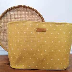 Op de afbeelding: Een mosterdgele draagtas met een wit sterrenpatroon. De tas heeft een rechthoekige vorm en een bruin leren label met de tekst "HANDMADE". Achter de tas staat een ronde rieten mand.