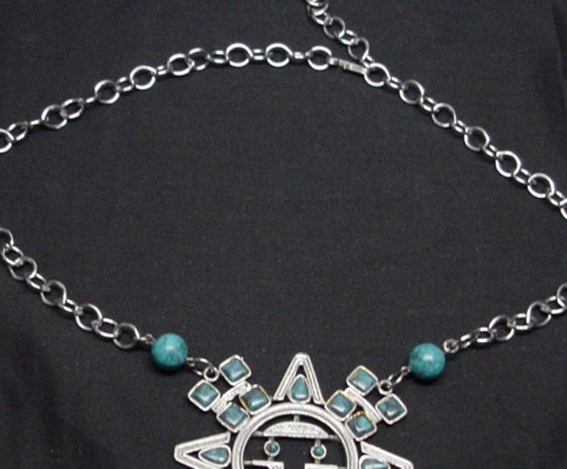 K&ouml;nnte beinhalten: Eine silberne Kettenkette mit einem t&uuml;rkisfarbenen Sonnenanh&auml;nger. Der Anh&auml;nger ist aus Metall und hat ein geometrisches Design. Die Kette ist silberfarben und hat ein einfaches Gliederdesign.