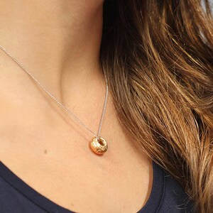 Puede incluir: Un collar de cadena de plata con un colgante de oro en forma de esfera. El colgante tiene una superficie texturizada.