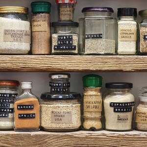 May include: A shelf filled with glass jars containing sand from different locations around the world. Each jar has a label with the location name and a small black tag with the country of origin. The jars are arranged in two rows on a wooden shelf.