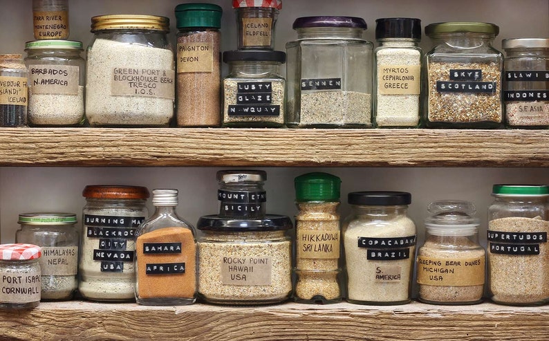 May include: A shelf filled with glass jars containing sand from different locations around the world. Each jar has a label with the location name and a small black tag with the country of origin. The jars are arranged in two rows on a wooden shelf.