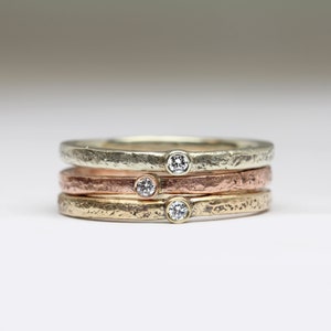 May include: Three thin gold bands with a textured finish. Each band has a small, clear diamond set in the center.