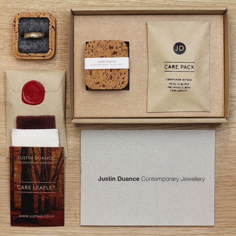May include: Overhead view of a jewellery presentation set. A gold ring in a cork box, a box with a cork coaster, a care pack, and a care leaflet are arranged on a wooden surface. The care pack contains a metal scourer and polishing cloth.