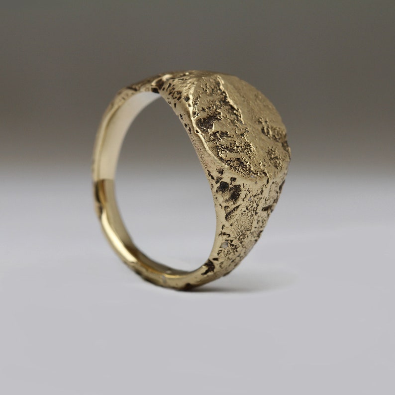 May include: A gold signet ring with a textured, irregular oval top. The ring has a smooth band.