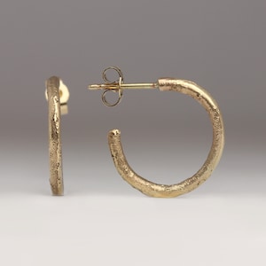 Puede incluir: Un par de pendientes de aro dorados. Los pendientes tienen una superficie texturizada y un cierre de poste y mariposa. Los aros tienen aproximadamente 2,5 cm de diámetro, mostrando un diseño minimalista.