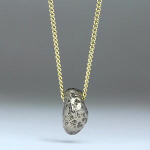 Puede incluir: Un collar con un colgante de plata de forma de roca, texturizado y rugoso, que cuelga de una cadena dorada.